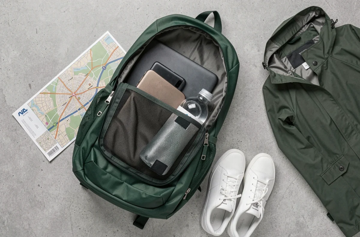 Flat-lay d'un sac à dos ville léger vert sapin ouvert avec ses accessoires sur fond béton gris